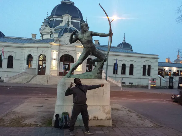 A traveller posing near an archer statue and a pale building