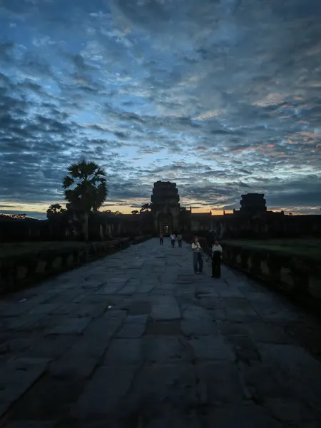 A stone causeway leading towards Angkor temple towers beneath a changing sky