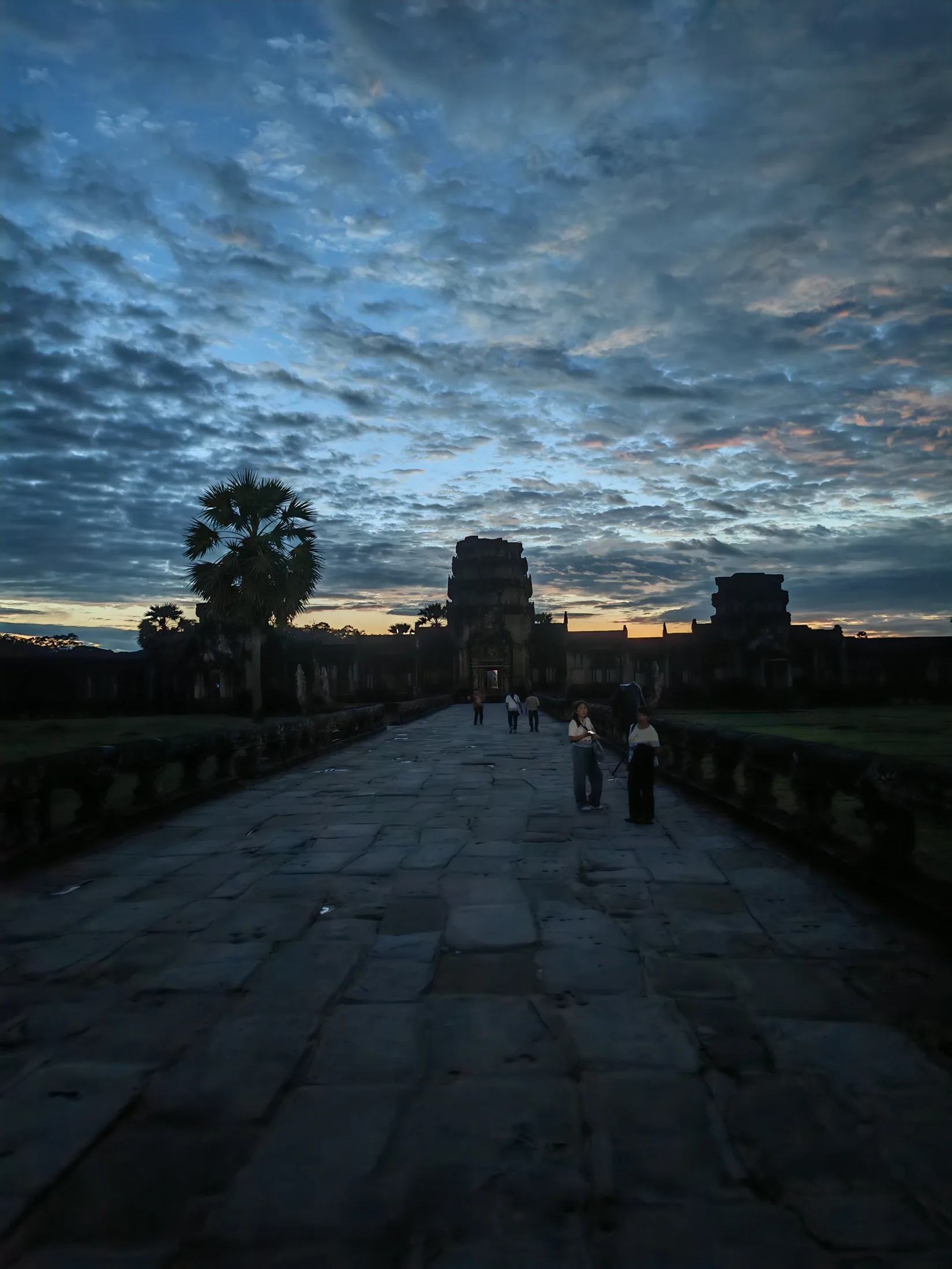 A stone causeway leading towards Angkor temple towers beneath a changing sky