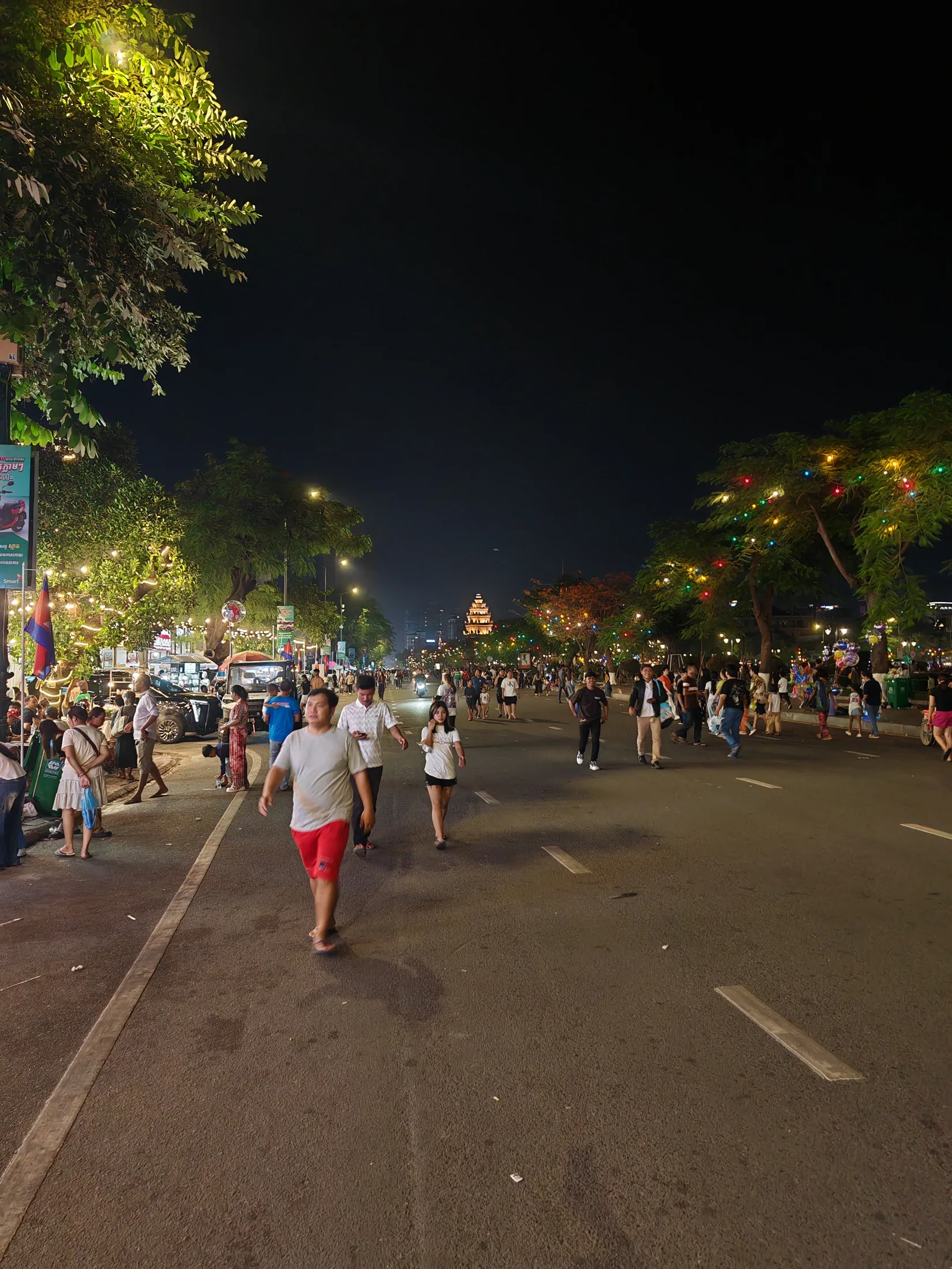 People walking along a brightly lit Phnom Penh boulevard at night