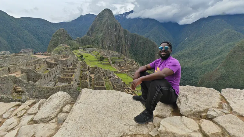 A male traveller seated on a stone wall overlooking Machu Picchu